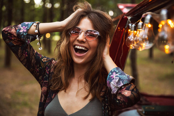 Image of young hippy woman wearing stylish accessories smiling while resting in forest camp - Photo, Image