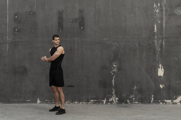 Strong muscular, sporty man warming up, getting ready for training, with a gray urban wall in background. Young sportsman stretching hands and triceps. Exercising and workout concept - Photo, Image