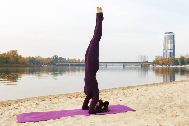 A young girl practices yoga by the river. Exercises against the background of the river, the shore and the bridge. - Photo, Image