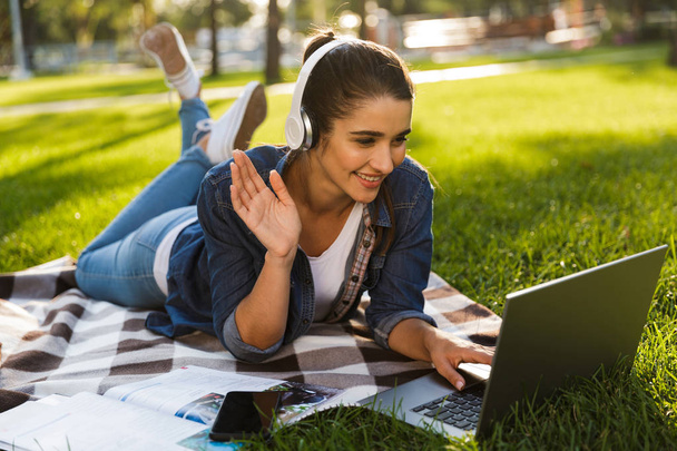Image of amazing happy woman student lies outdoors in a park using laptop computer listening music waving. - Zdjęcie, obraz