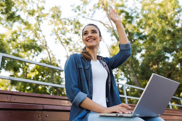 Image of amazing young beautiful woman sitting outdoors using laptop computer waving. - Foto, afbeelding