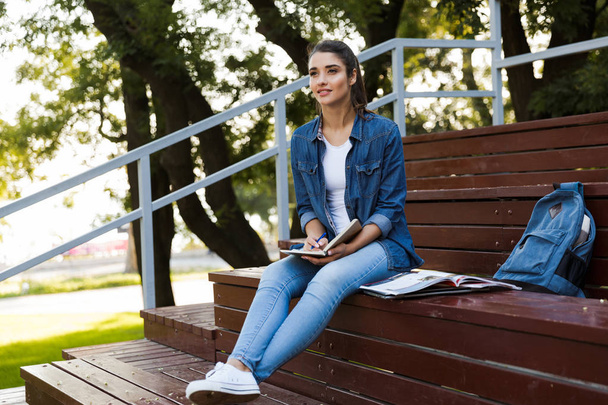 Image of amazing young beautiful woman sitting outdoors writing notes in notebook. - Valokuva, kuva