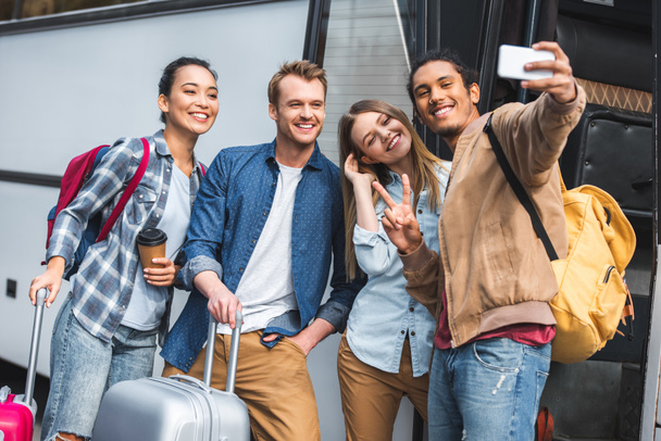 wesoły wielorasowe turystycznych Wyświetlono znak pokoju i biorąc selfie z wielokulturowym znajomych na smartfon w pobliżu podróży autobusem na ulicy - Zdjęcie, obraz