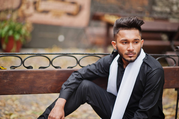 Indian stylish man in black traditional clothes with white scarf posed outdoor sitting on bench. - Fotó, kép