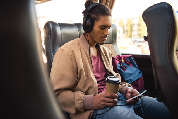 multiracial male tourist in headphones sitting with paper coffee cup and using smartphone in bus - Photo, Image
