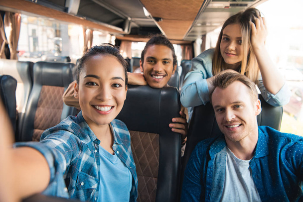 selective focus of smiling asian woman taking selfie with multicultural friends in travel bus  - Photo, Image