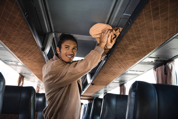 sorrindo jovem multirracial homem colocando mochila na prateleira em ônibus de viagem
  - Foto, Imagem