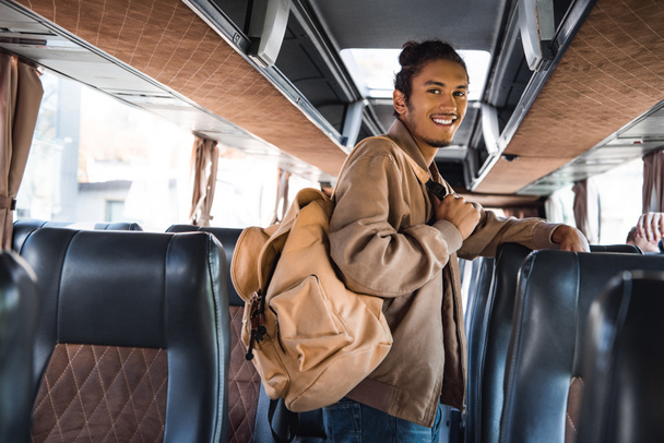 sonriente turista masculino multirracial con mochila mirando a la cámara en autobús de viaje
 - Foto, Imagen