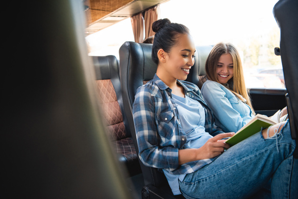 Смеющиеся многонациональные подруги читают книги во время поездки на туристическом автобусе
 - Фото, изображение
