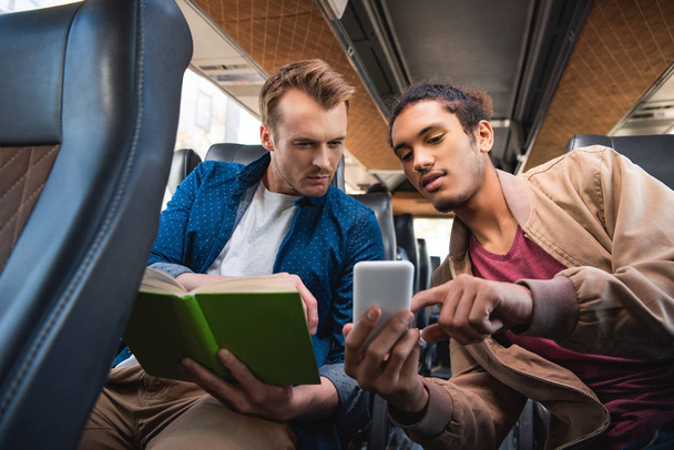 young mixed race man showing smartphone to his male friend with book during trip on travel bus - Photo, image