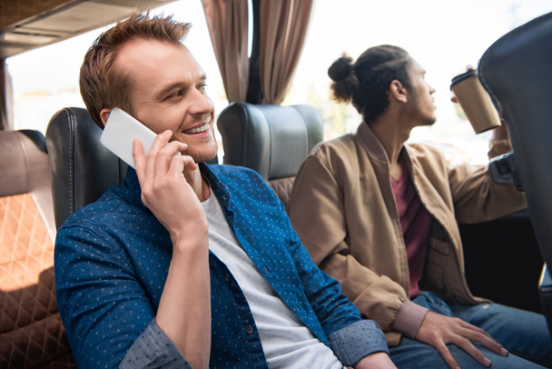 улыбающийся мужчина разговаривает на смартфоне в то время как его друг пьет кофе рядом во время поездки на автобусе
 - Фото, изображение