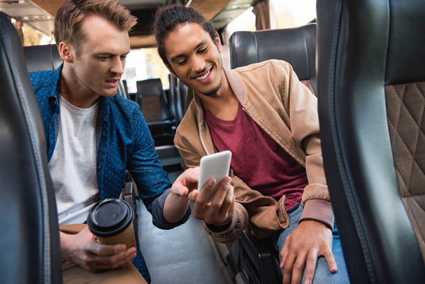 felice uomo di razza mista mostrando smartphone al suo amico maschio con tazza di caffè usa e getta durante il viaggio in autobus
 - Foto, immagini