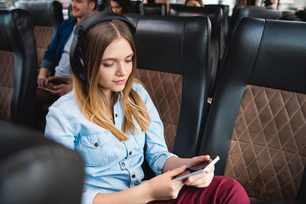 young woman in headphones listening music and using smartphone during trip on travel bus - Foto, afbeelding