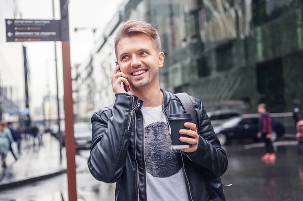 Attractive stylish young man in a leather jacket with a smartphone and take-out coffee in his hands against the backdrop of a large modern city - Photo, image