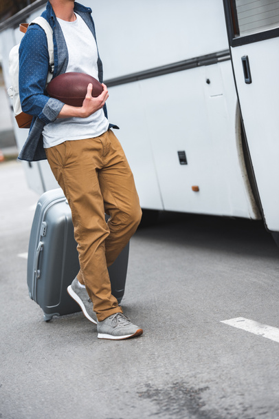 обрізане зображення людини з рюкзаком і м'ячем регбі, що перевозить сумку на колесах поблизу туристичного автобуса на вулиці
  - Фото, зображення
