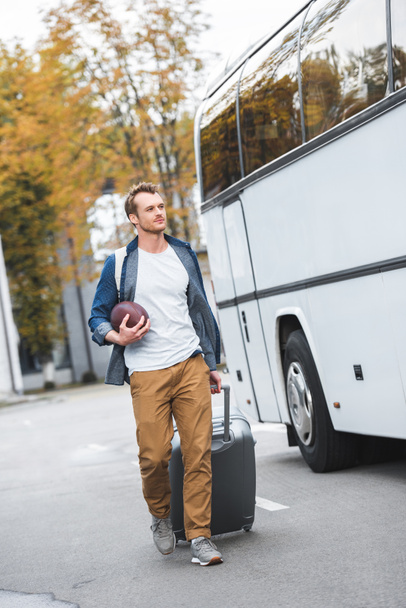 homem bonito com mochila e bola de rugby levando saco sobre rodas perto de ônibus de viagem na rua
  - Foto, Imagem