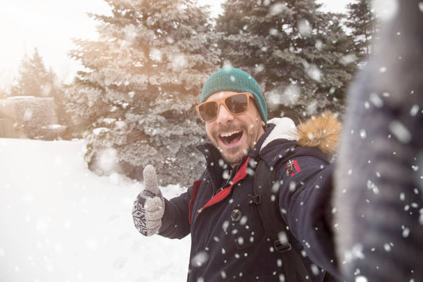Uomo con gli occhiali da sole prendere selfie il giorno nevoso e mostrando pollice in su
. - Foto, immagini