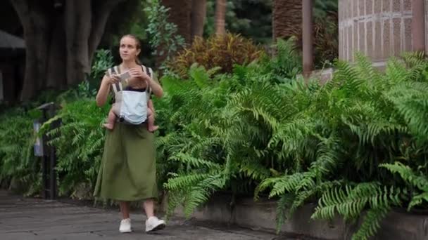 Женщина в платье с ребенком в стропе идет вдоль пальм и снимает видео на мобильный телефон
. - Кадры, видео