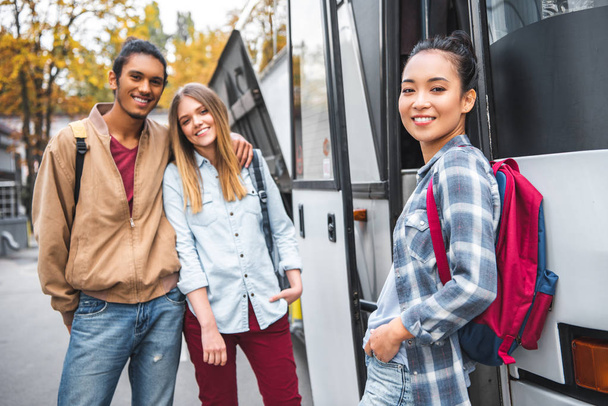 junge multiethnische Freunde mit Rucksäcken, die in der Nähe eines Reisebusses auf der Straße in die Kamera schauen  - Foto, Bild
