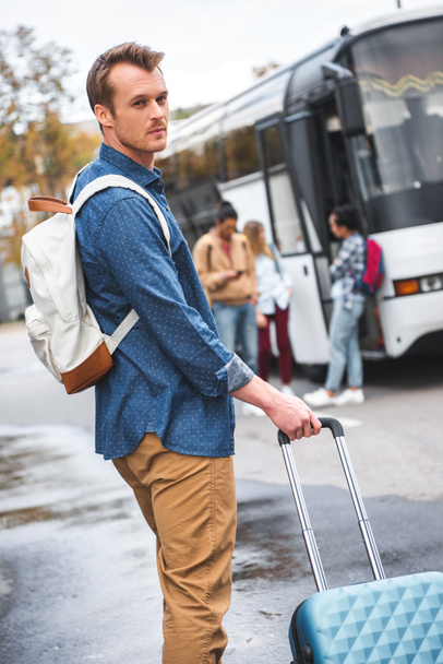 selective focus of handsome man with backpack carrying wheeled bag near travel bus at street - 写真・画像
