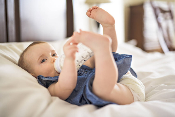 Pretty baby girl drinks water from bottle lying on bed - Foto, Bild