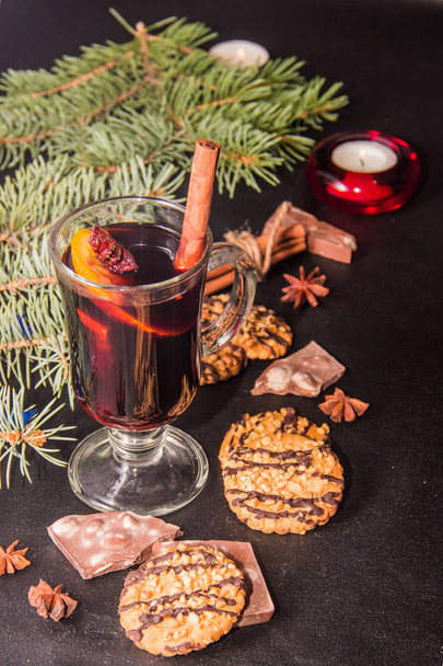 Una copa de vino caliente con sabor sobre un fondo de cuero negro con trozos de galletas de palillos de chocolate y una rama de árbol de Navidad
 - Foto, Imagen