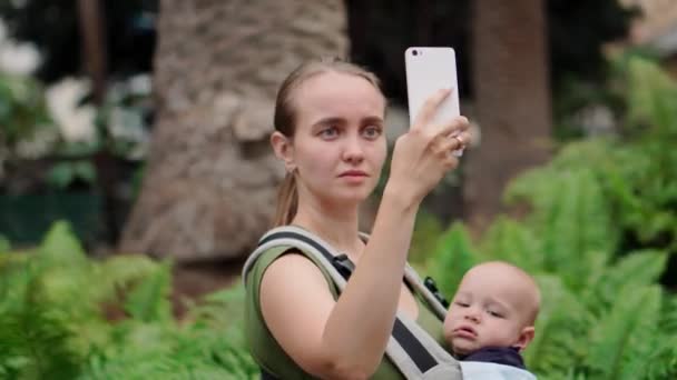 Мама с ребенком в стропе держит в руках смартфон и общается через видеосвязь с друзьями
 - Кадры, видео