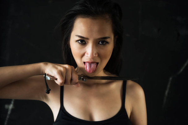 Portrait of psychosis sadism woman holding knife at her neck and show tongue to threaten a victim with dark black background and copy space for text. Halloween, Crime, Drug, Abuse for safety concept. - Photo, Image