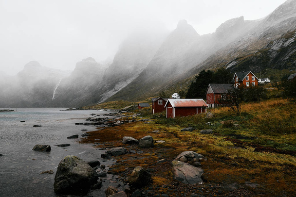 Pueblo abandonado Vindstad en Noruega en las islas Lofoten con casas de madera roja en primer plano y montañas y el océano en el fondo
.       - Foto, Imagen