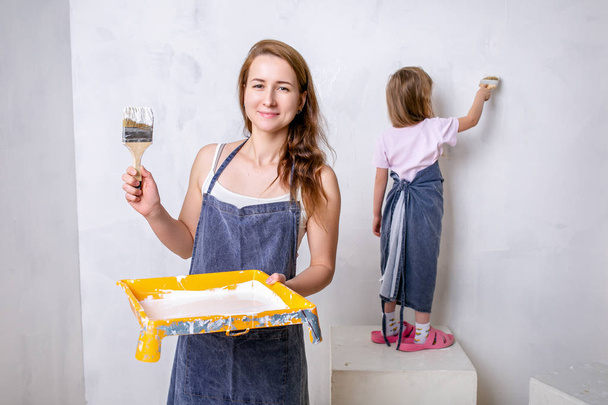 Repair in the apartment. Happy family mother and little daughter in blue aprons paints the wall with white paint. They paint the wall with brushes and smiling. Horizontal portrait - Photo, image
