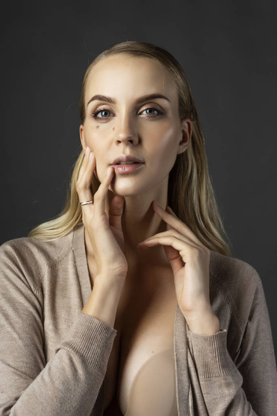 A beautiful blonde girl model wearing a knitted beige dress, unbuttoned on her breasts and a bra touches her face with her hand. Nude makeup. Clean, healthy skin. Gray background. Copy space - Φωτογραφία, εικόνα