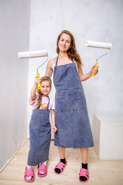 Repair in the apartment. Happy family mother and little daughter in blue aprons paints the wall with white paint. Mom and daughter are holding paint rollers and smiling. Horizontal portrait - 写真・画像