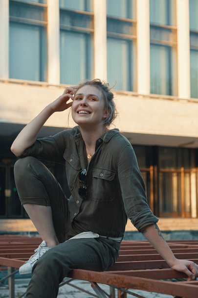 Portret van een casual blond meisje zittend op de metalen constructie met een gebouw op de achtergrond. Jonge moderne vrouw poseren in de stad, buiten. Stadsleven - Foto, afbeelding