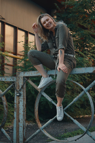 Chica elegante posando en la calle sentado en una valla de acero con las piernas dobladas. Estilo urbano. Moda callejera
. - Foto, imagen