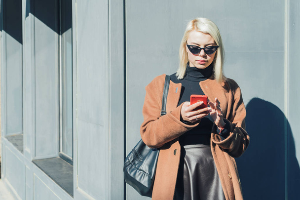 Portrait of young attractive blonde woman using mobile device in autumn city. Girl have stylish look, sunglasses and nose piercing. Technology concept. Copy space - Photo, Image