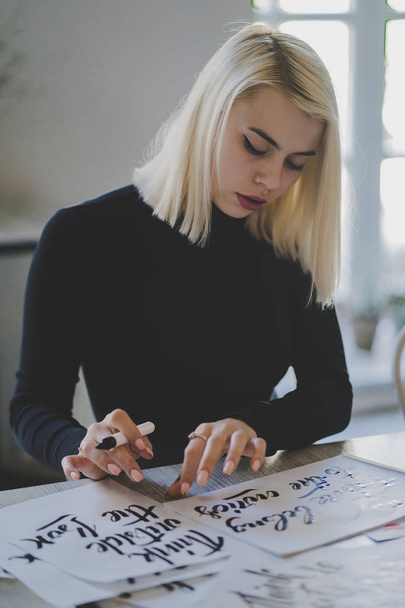 Portrait of young girl sitting in library or office and working with lettering on papers. Blonde woman with perfect makeup and short hairstyle. Education, business concept. - Photo, image