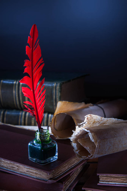 Red quill pen and a rolled papyrus sheet on a wooden table with old books - Foto, Bild