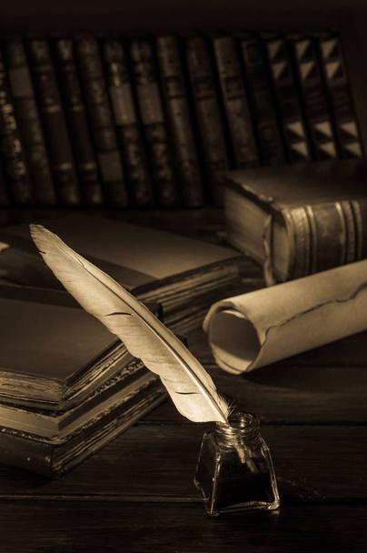 Pluma de pluma y una hoja de papiro enrollado en una mesa de madera con libros antiguos, efecto sepia
 - Foto, Imagen
