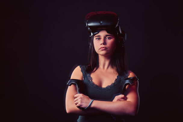 Portrait d'une jeune femme portant des lunettes de réalité virtuelle et tenant des joysticks, posant avec les bras croisés, regardant la caméra
. - Photo, image