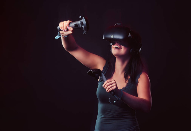 Beautiful young woman with virtual reality headset and joysticks playing video games. - Photo, Image