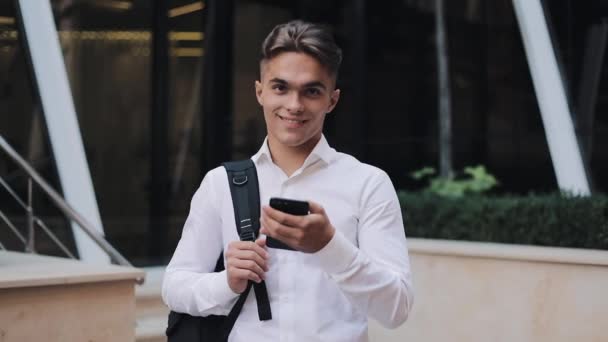 Handsome young businessman or tourist holding a phone in hands, looking into the camera and smiling. Concept: new business, travel the world, communication, contacts, communications, business deal - Footage, Video