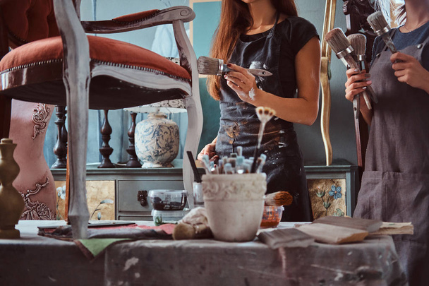 Artista femenina con su asistente en delantales pintando silla vintage con pincel en taller
. - Foto, imagen