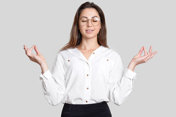 Photo of peaceful calm young woman holds hands in mudra gesture, tries to relax and concentrate before important business meeting, dressed in formal clothes, spectacles, isolated on white wall - Photo, image