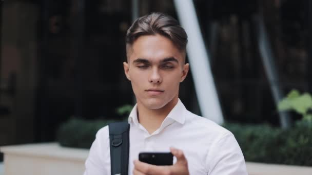 Handsome young businessman or tourist holding a phone in hands, looking into the camera and smiling. Concept: new business, travel the world, communication, contacts, communications, business deal - Imágenes, Vídeo