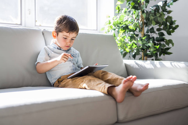 Dijital tabletli küçük çocuk kanepede oturuyor, evin arka planında. - Fotoğraf, Görsel