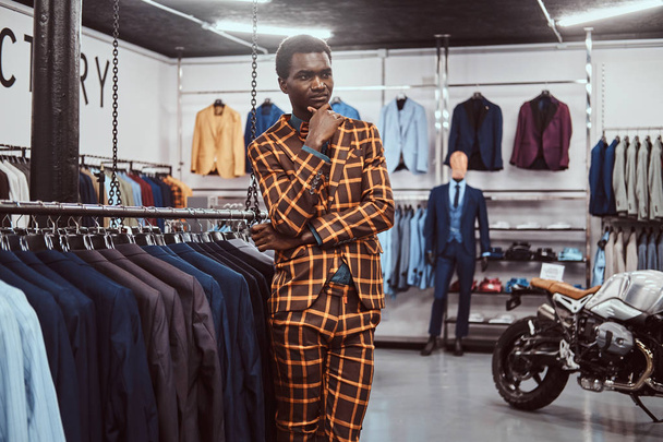 Elegantemente vestito uomo africano in posa con mano sul mento mentre in piedi in un negozio di abbigliamento maschile classico
. - Foto, immagini