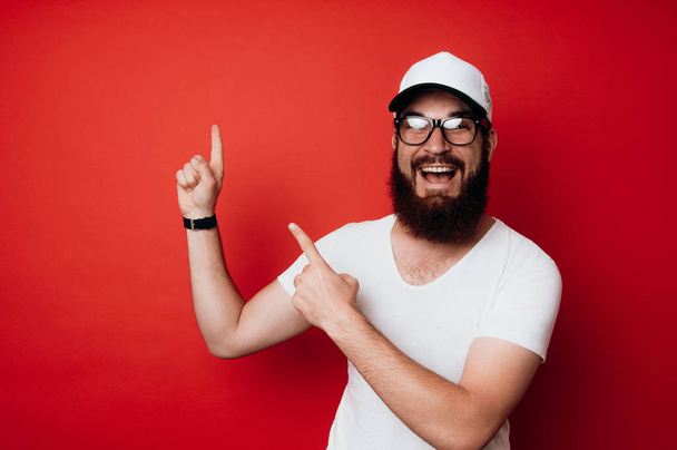 Cheerful bearded hipster man wearing eyeglasses and pointing at copyspace over red background - Foto, Bild