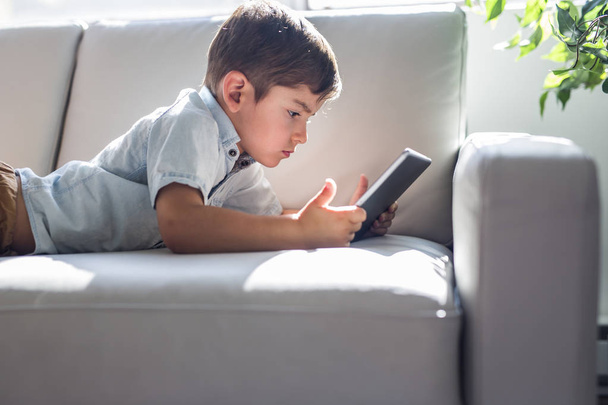Little boy with digital tablet sitting on sofa, on home interior background - Zdjęcie, obraz