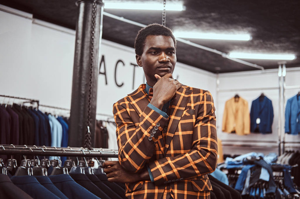 Элегантно одетый африканский мужчина позирует с рукой на подбородке, стоя в классическом магазине мужской одежды
. - Фото, изображение
