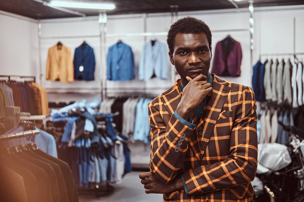 Elegantly dressed African man posing with hand on chin while standing in a classic menswear store. - Фото, зображення